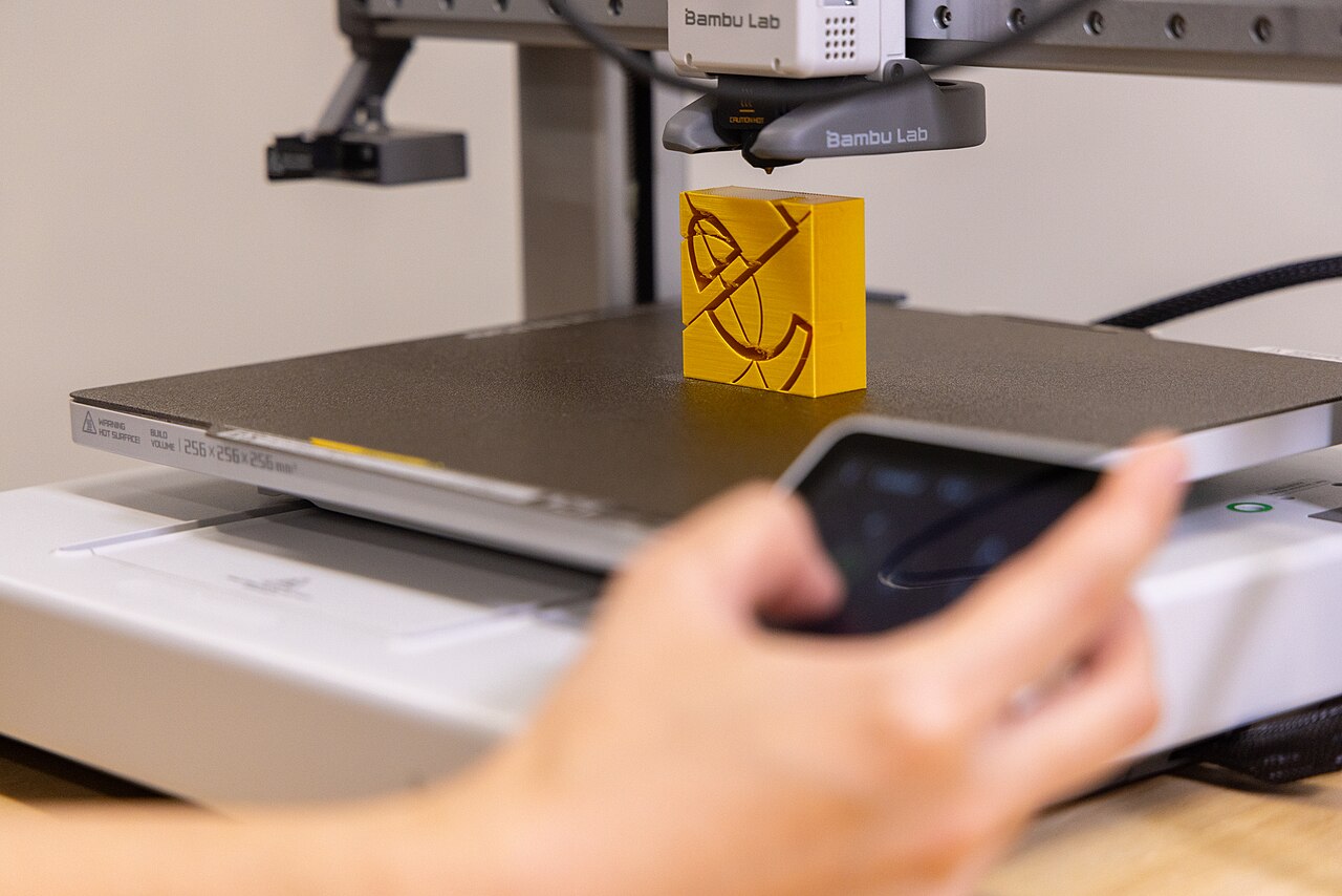 A yellow printed piece on a Bambu Lab A1 build plate, with the toolhead above it.